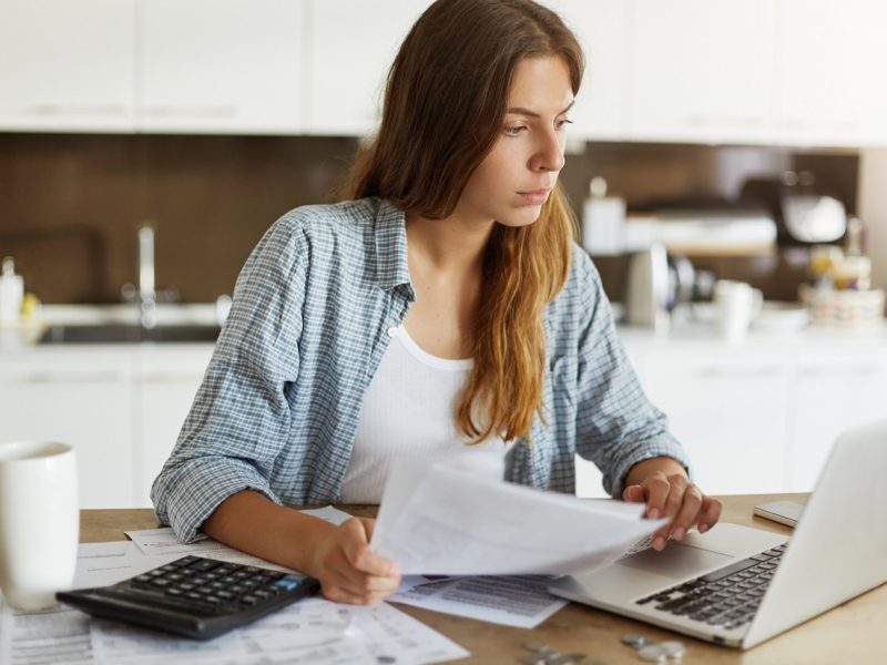 Indoor portrait of concentrated young European female sitting at kitchen table, using banking application on laptop computer while paying electricity and gas bills online. Family budget and finances