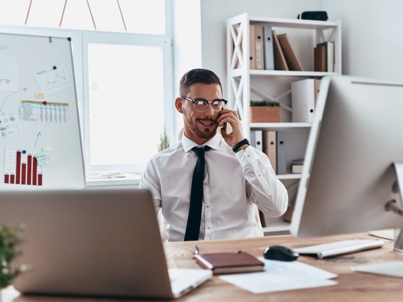 Talking with client. Handsome young man in formalwear talking on the phone and smiling while sitting in the office